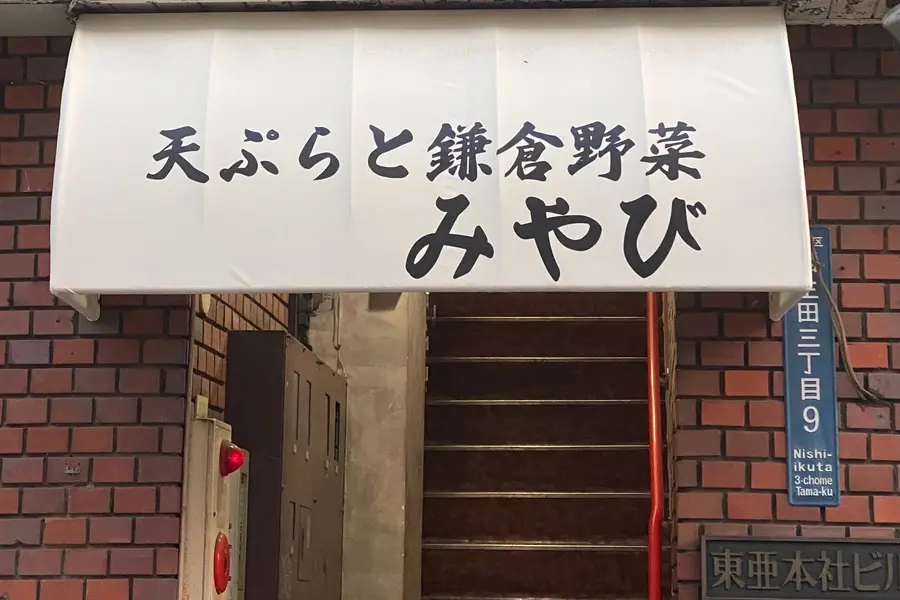 天ぷらと鎌倉野菜 みやびの外観。白い日除けに店名、東亜本社ビル2階へ続く階段